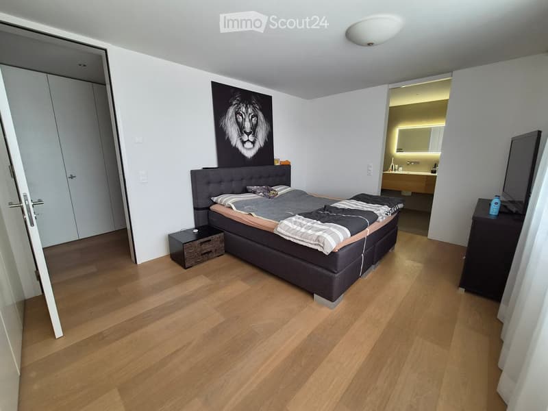 Modern bedroom with wooden flooring, a large bed with grey bedding, a nightstand, a mirror, and a picture of a lion on the wall.