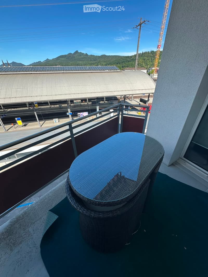 Balcony with a round wicker table and a metal railing, overlooking a train station and distant mountains.