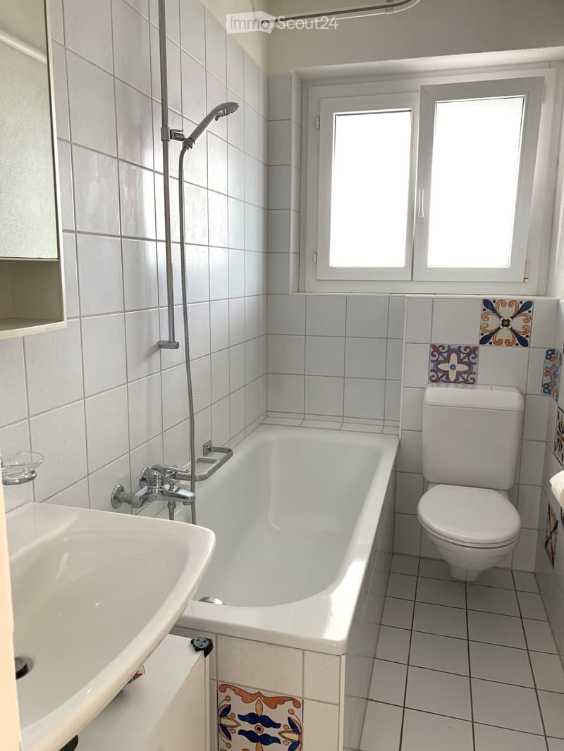 Modern bathroom with white tiles, a bathtub, toilet, sink, and a showerhead. Features decorative tiles on the walls and a window for natural light.