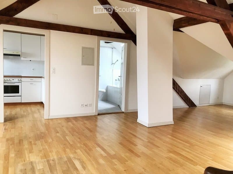 Bright, open loft-style room with wooden flooring, white walls, exposed wooden beams, and visible kitchen and bathroom areas. The space is completely unfurnished.