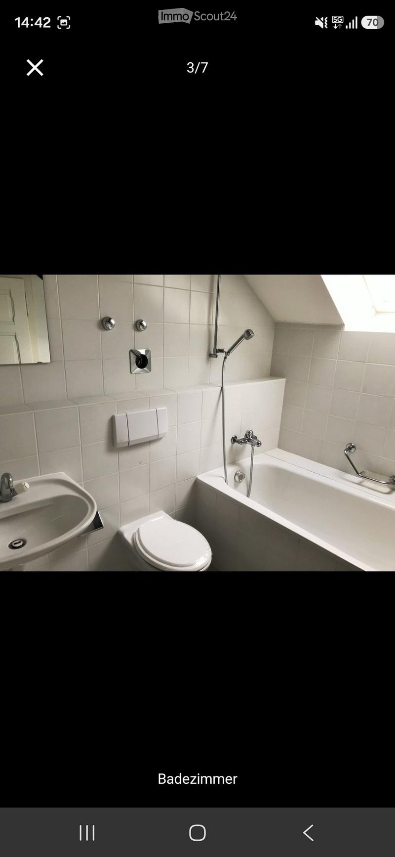 Modern bathroom with white tiles, a bathtub, toilet, sink, and mirror. Features a handheld showerhead and a window for natural light.