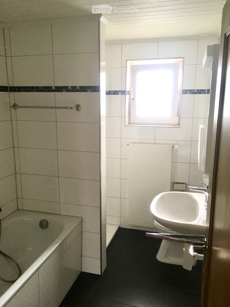Modern bathroom with white tiles, bathtub, sink, toilet, and a window providing natural light.
