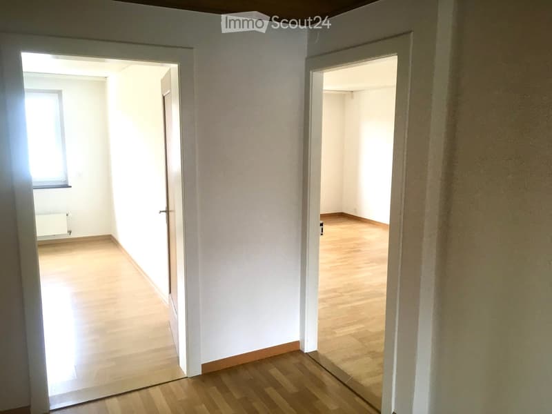 Empty hallway with wooden flooring and white walls, leading to two open rooms with similar flooring and walls.