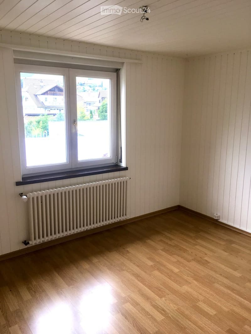 Empty room with wooden flooring, white paneled walls, a radiator, and a large window with a view of neighboring houses.