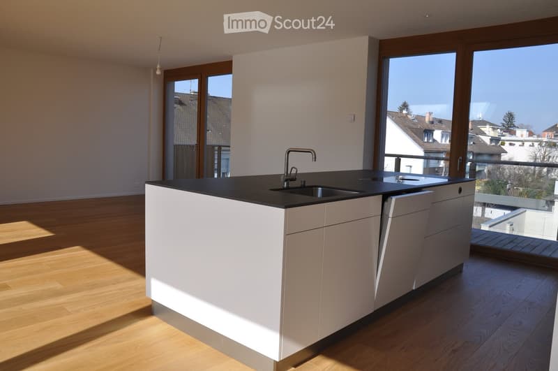 Modern kitchen with white cabinets, black countertop, and a central island featuring a sink and faucet. Large windows provide natural light and a view of the neighborhood.