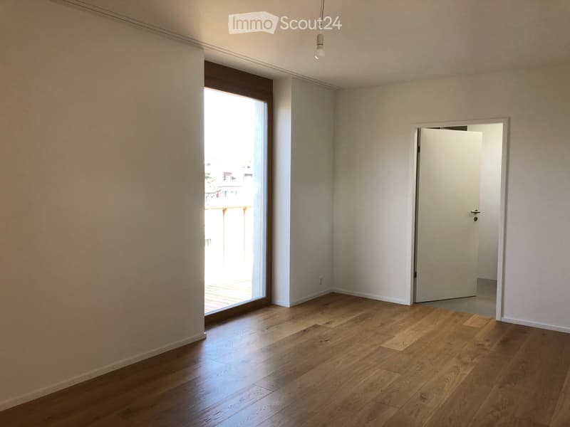 Empty room with wooden flooring, white walls, large window with view, and a closed door leading to another space.
