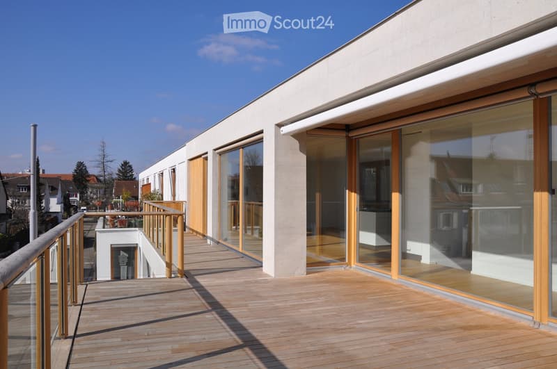 Modern wooden deck terrace with glass sliding doors, wooden railing, and a view of neighboring buildings and trees.