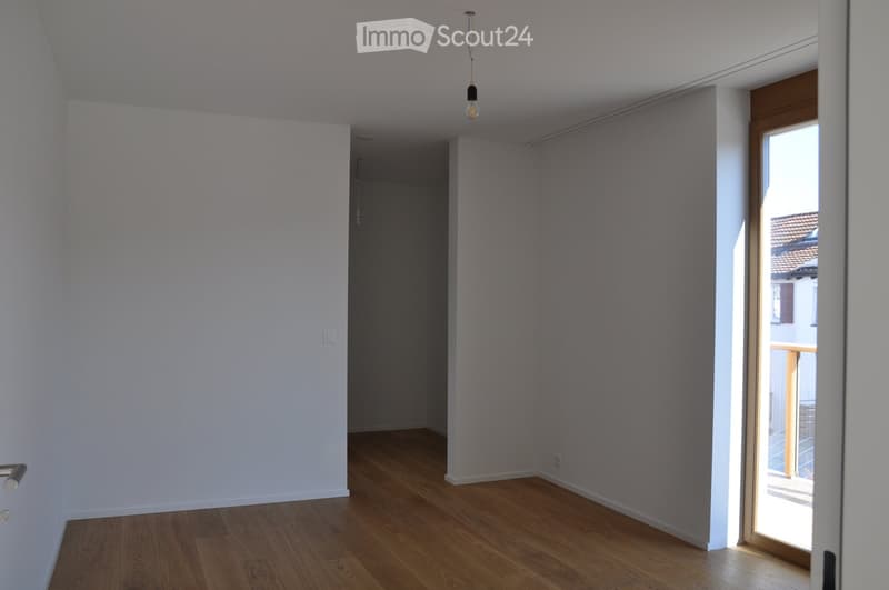 Empty room with light-colored walls and wooden flooring, featuring a pendant light fixture and an open doorway leading to a balcony area.