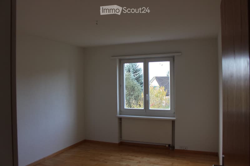 Empty room with wooden flooring, white walls, and a large window with a view of trees and a neighboring house.