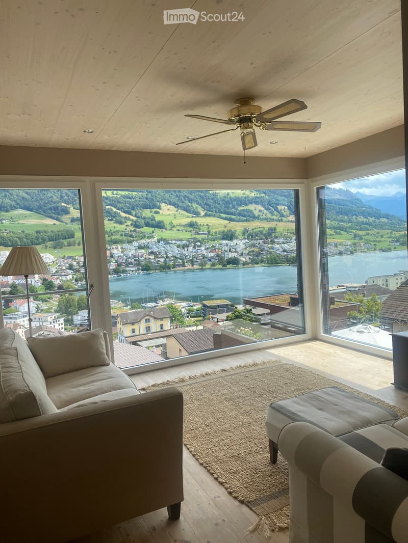 Modern living room with large windows offering a scenic view of a lake and surrounding landscape, featuring a sofa, ottoman, floor lamp, and ceiling fan.
