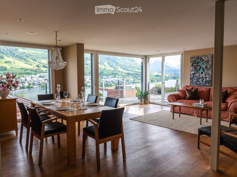 Elegant living and dining area with wooden flooring, large windows offering scenic views, a red patterned sofa, a wooden dining table with chairs, and decorative items including a flower vase and candles.