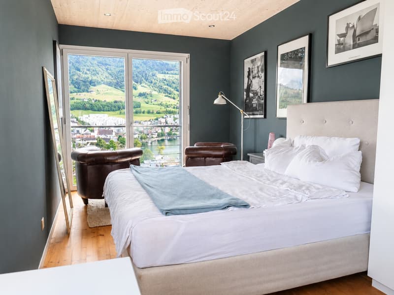Modern bedroom with a large bed, two leather chairs, a floor lamp, and a scenic view through large windows.