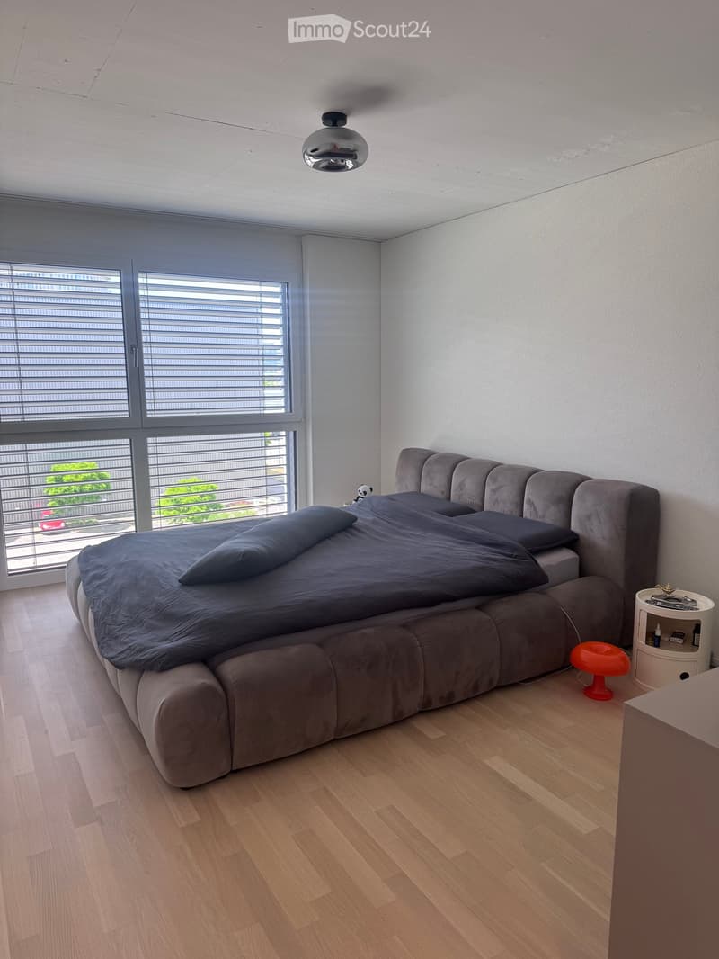 Modern bedroom with a large grey sofa bed, dark blue bedding, a small nightstand with a table lamp, and a red stool. Large windows with blinds offer a view of the city.
