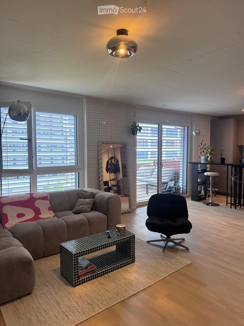 Modern living room with a grey sectional sofa, coffee table, black chair, pendant light, floor lamp, and a view of a balcony area with outdoor furniture.