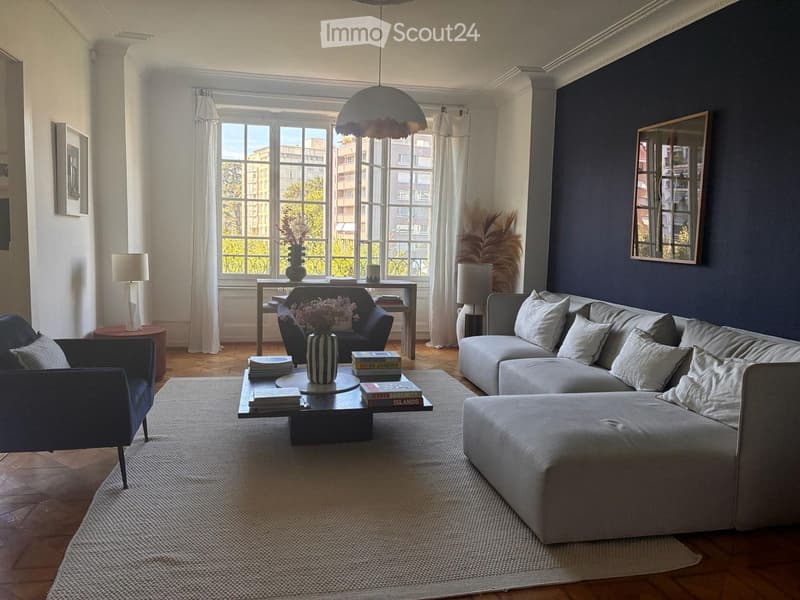 Modern living room with large windows, dark blue accent wall, grey sectional sofa, coffee table with books and vase, blue armchair, and decorative lamps.