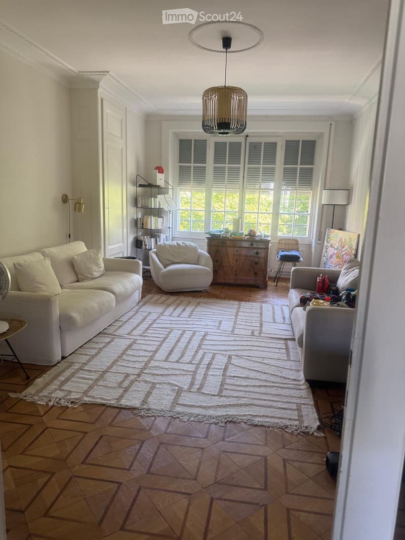 Bright living room with wooden flooring, large windows, and natural light. Features a cream-colored sectional sofa, armchair, coffee table, side table, and a woven area rug.