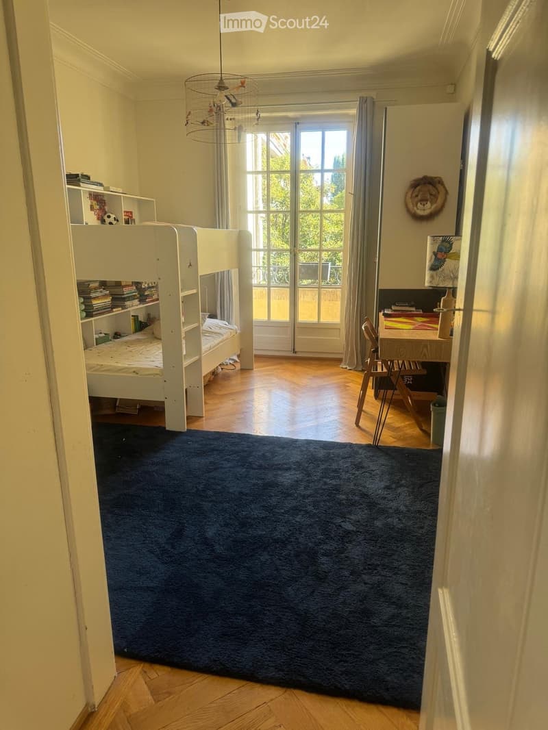 Bright bedroom with wooden flooring, large window, bunk bed, desk, and shelves.