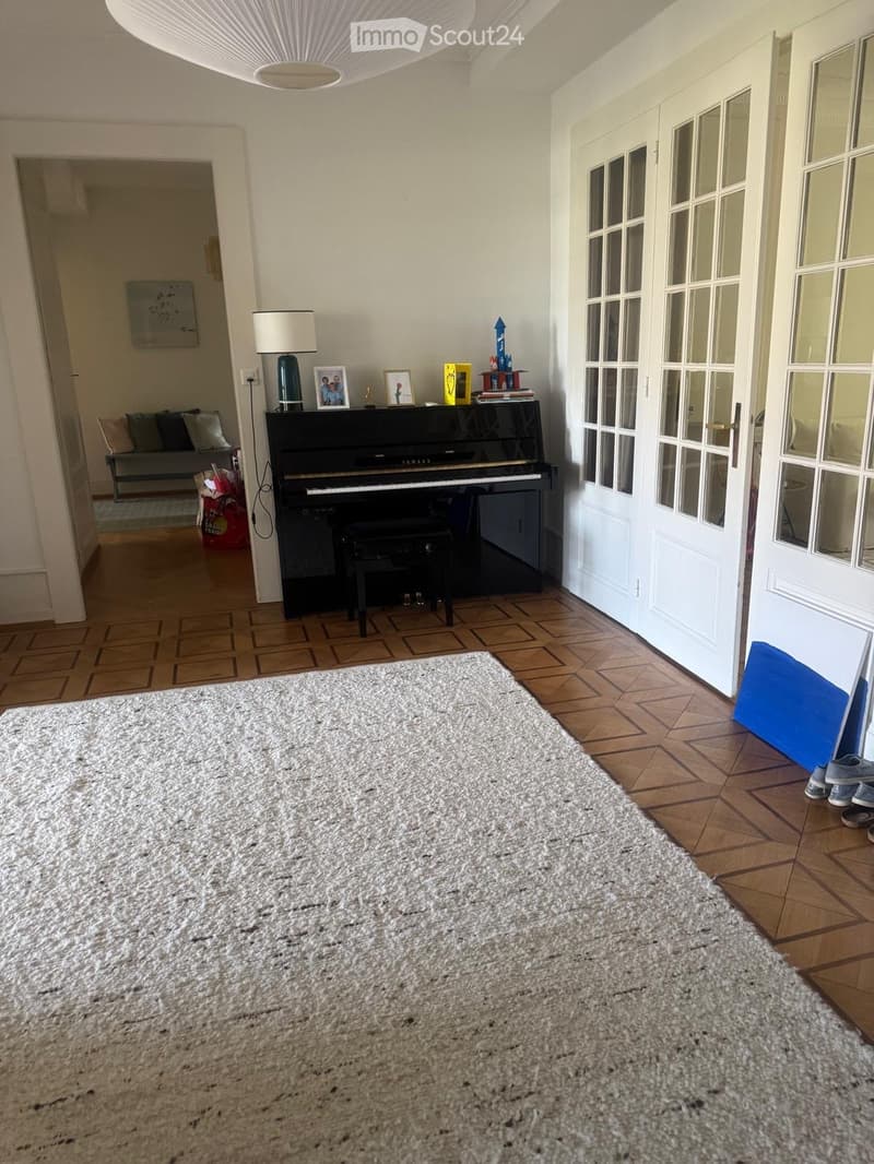 A well-lit room with wooden flooring, a large white rug, a black piano with a stool, a table lamp, picture frames, and a sofa visible in the background.