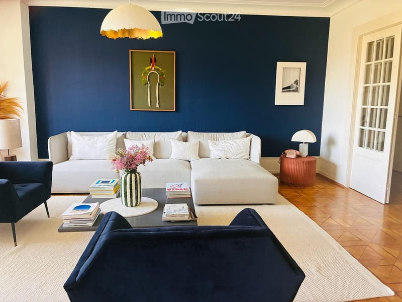 Modern living room with dark blue accent wall, white sectional sofa, coffee table with books and vase, armchair, and decorative lighting.