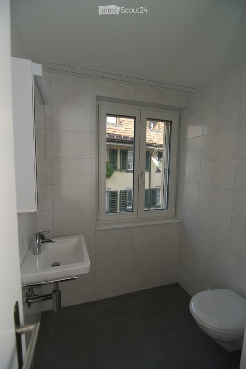 Modern bathroom with a white sink, chrome faucet, and toilet. The room features tiled walls and a window offering a view of a neighboring building.