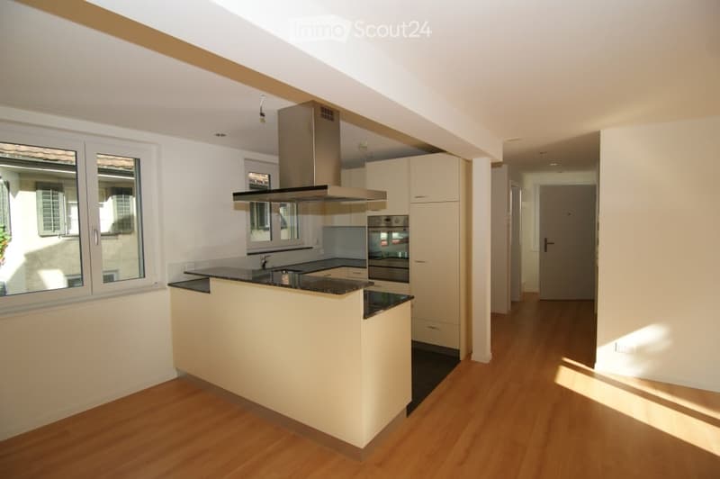 Modern kitchen with light-colored cabinets, a central island, and built-in appliances including an oven and range hood. The space features wooden flooring and large windows allowing natural light.