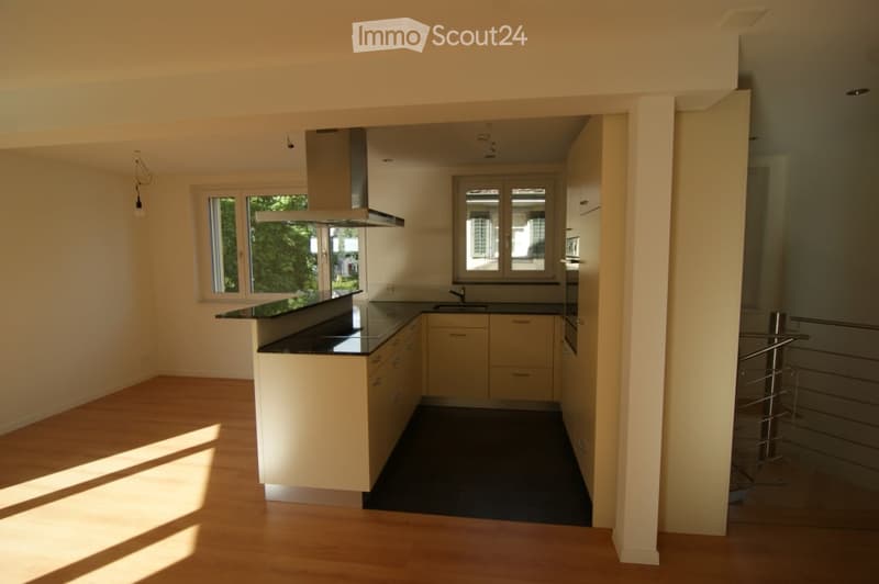 Modern kitchen with light-colored cabinets, black countertops, a central island, and a range hood. The space features wooden flooring and large windows allowing natural light to fill the room.