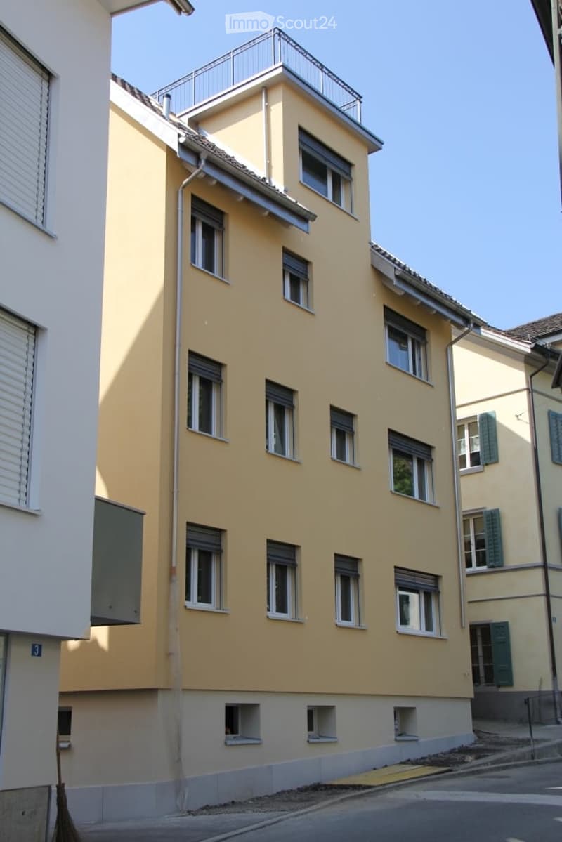Four-story residential building with yellow facade, multiple windows, and a small rooftop terrace with a metal railing.