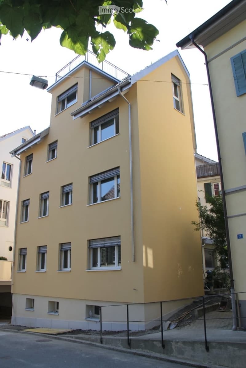 Four-story yellow building with multiple windows and a raised ground floor, surrounded by neighboring structures and greenery.