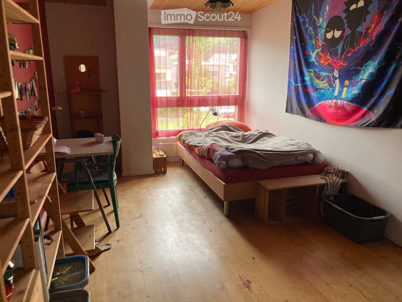 Cozy bedroom with wooden floor, bed with red mattress, small table with chair, shelf with books and basket, and a colorful tapestry on the wall.