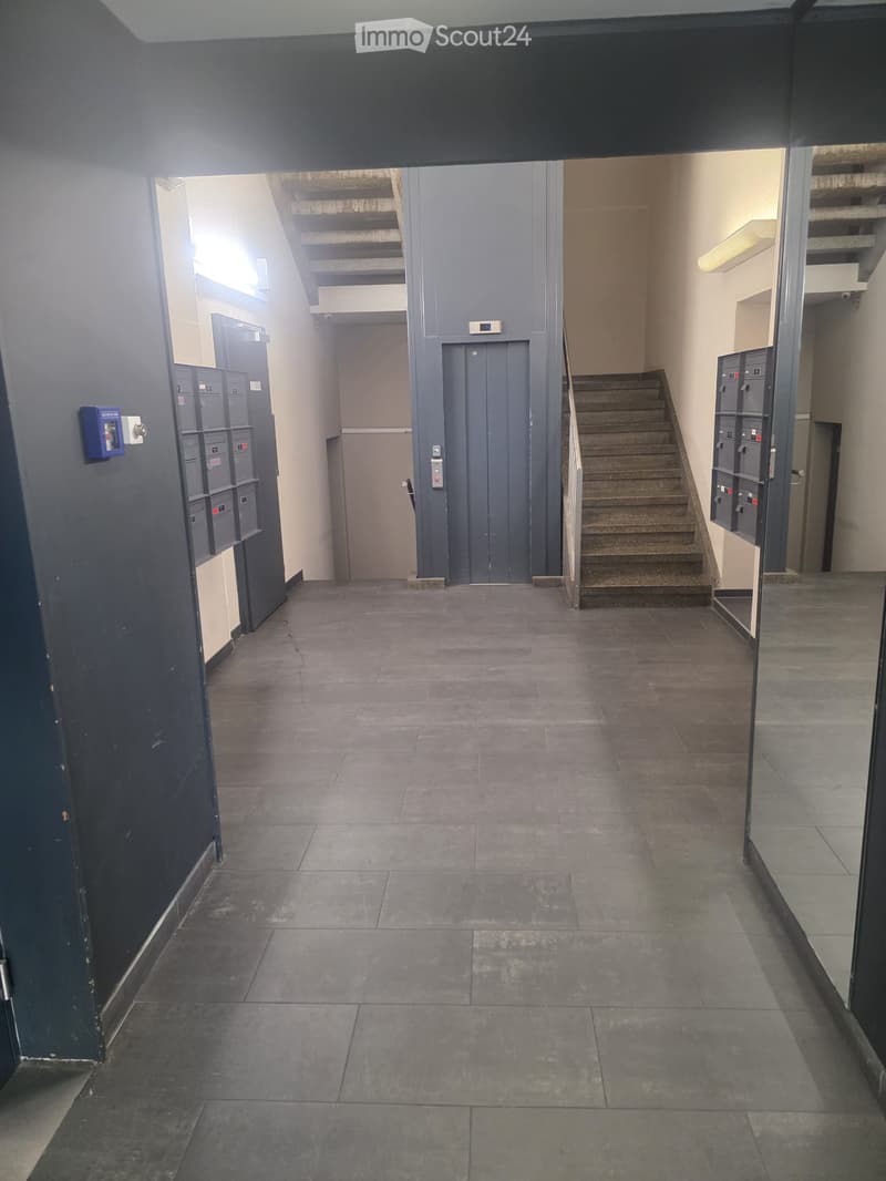 Empty hallway with tiled floor, staircase leading upwards, elevator, and mailboxes on both sides.