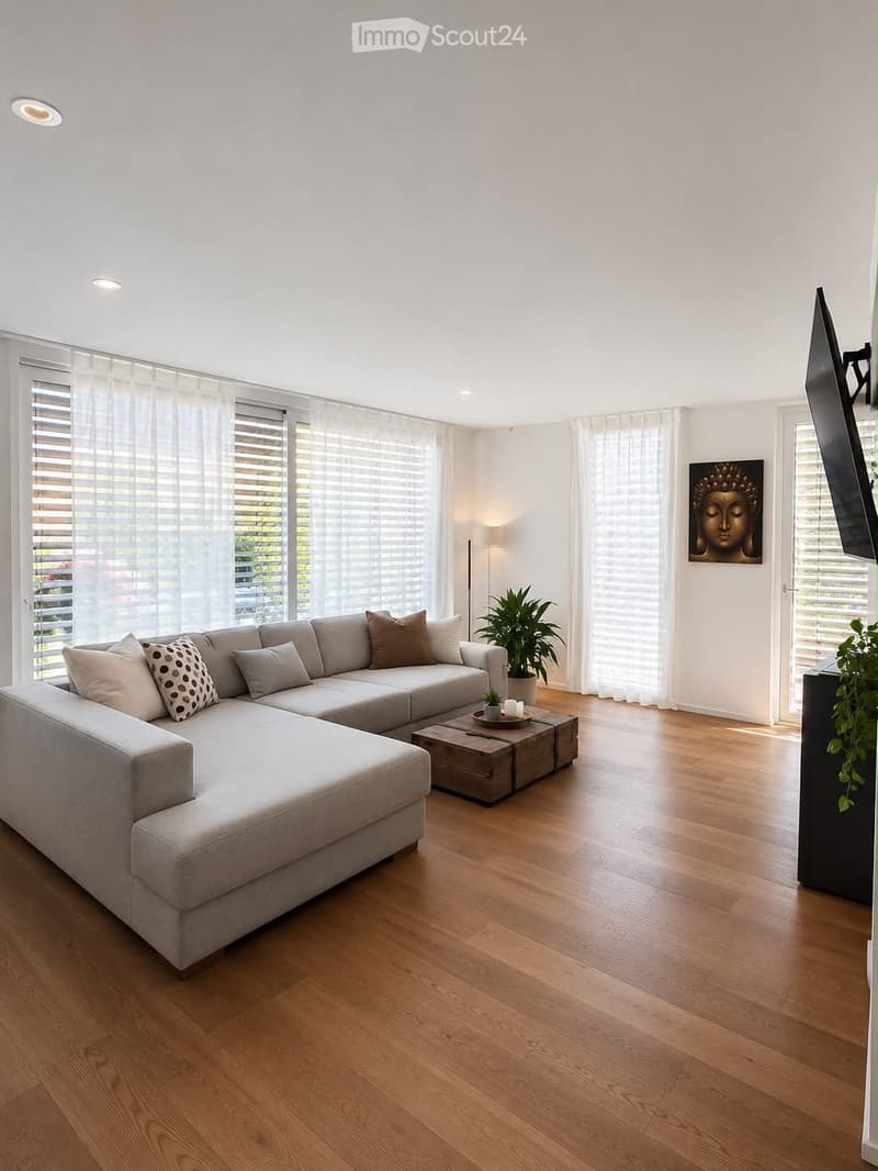 Modern living room with light wooden flooring, large windows with blinds, a grey sectional sofa, a coffee table, a floor lamp, and a television mounted on the wall.