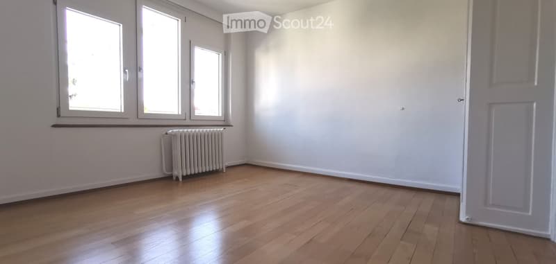 Empty room with wooden flooring, white walls, and three windows. A radiator is visible under the windows on the left side.