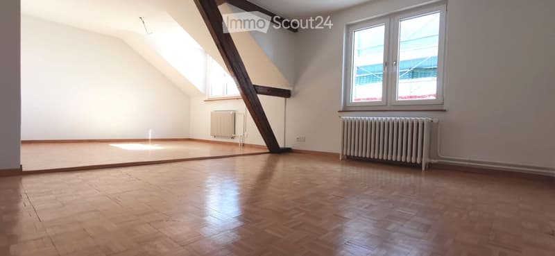 Empty attic room with wooden flooring, white walls, and large windows allowing natural light. Radiators are present along the walls.
