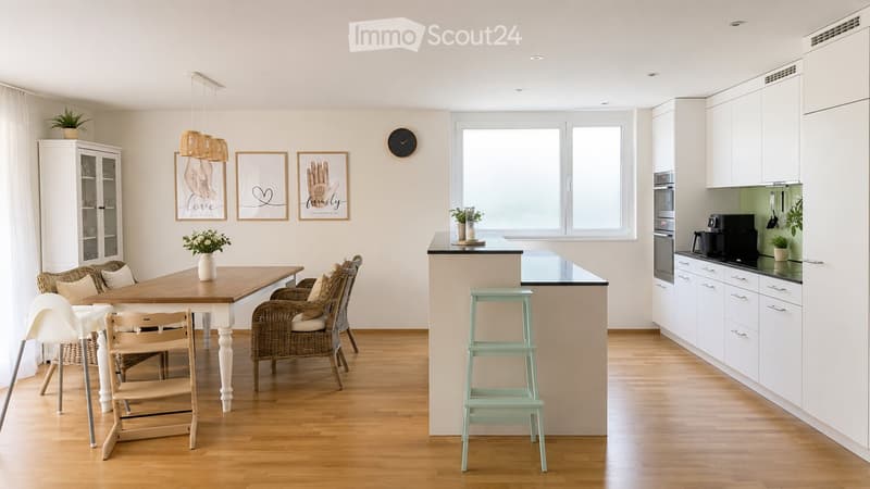 Bright, modern kitchen with white cabinets, a wooden dining table, and a small seating area. Features include a built-in oven, coffee maker, and decorative elements like framed art and potted plants.