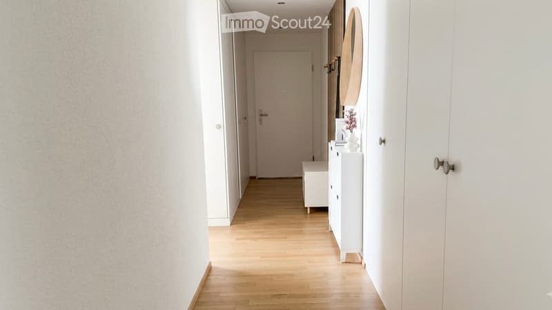 A clean, modern hallway with light wooden flooring, white walls, and a closed door at the end. A small white bench and a small white cabinet with a vase and mirror are visible on the right side.