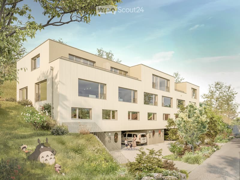 Modern multi-story residential building with large windows, balconies, and a raised ground floor entrance. The building is surrounded by greenery and has a paved pathway leading to the entrance.