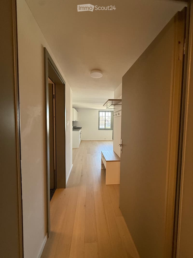 A narrow hallway with wooden flooring, white walls, and a bench on the right side. The hallway leads to a kitchen area with white cabinets and a window.
