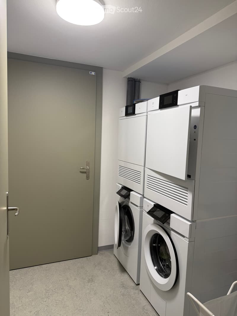 A small, well-lit room with a washing machine and dryer stacked side by side, featuring a grey door and a round ceiling light.