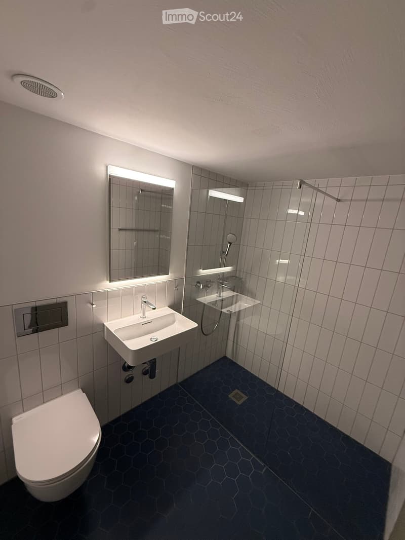 Modern bathroom with hexagonal dark blue tiles, white sink, toilet, and illuminated mirror above the sink.
