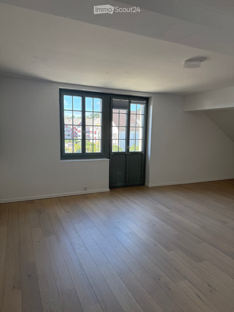 Empty room with light wooden flooring, grey walls, and large windows with grey frames, allowing natural light to enter.