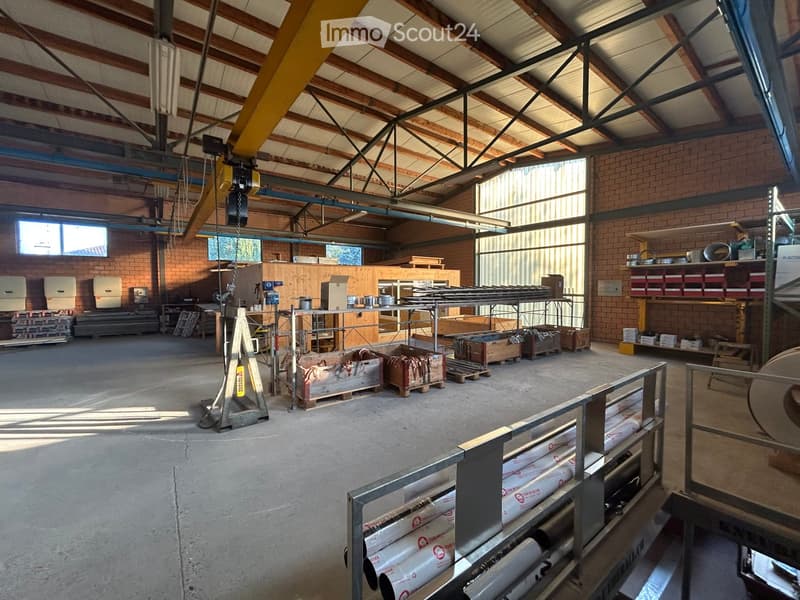 Empty warehouse space with exposed wooden beams, brick walls, large windows, and scattered storage items and materials.