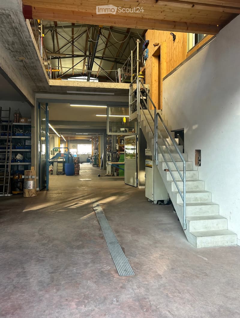 Empty industrial-style room with concrete floors, exposed wooden beams, and a staircase leading to an upper level. The space includes a large open area with scattered storage items and equipment in the background.