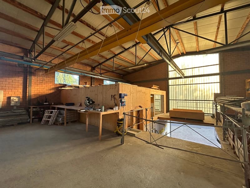 Empty workshop or garage space with exposed wooden beams, brick walls, large windows, and a concrete floor. No furniture or equipment is present.