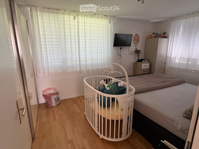 A well-lit bedroom with a double bed, a crib, a nightstand, and a television mounted on the wall. The room features wooden flooring and large windows with curtains.