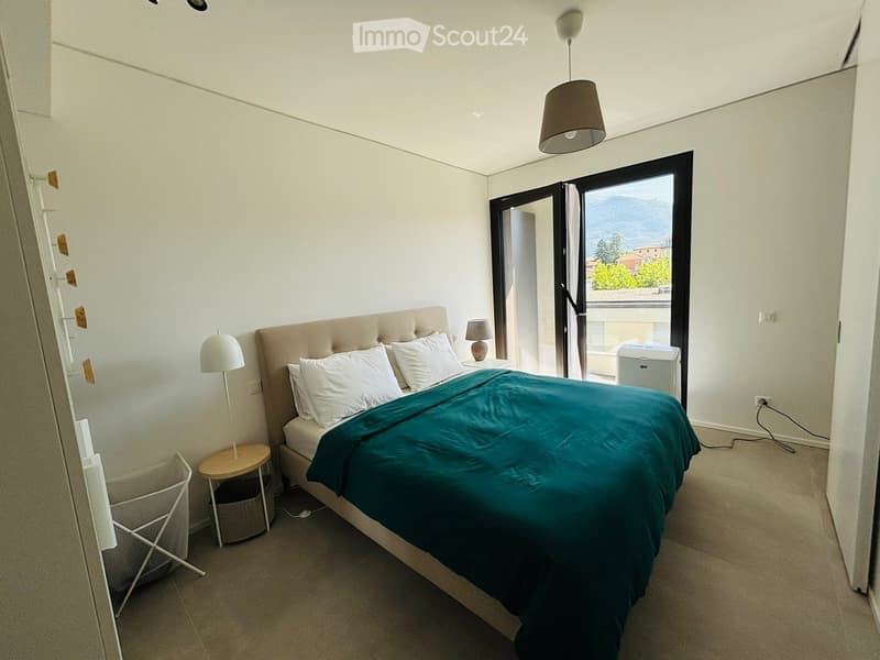 Modern bedroom with a large bed, nightstand, side table, and two lamps. Large window with a scenic mountain view.