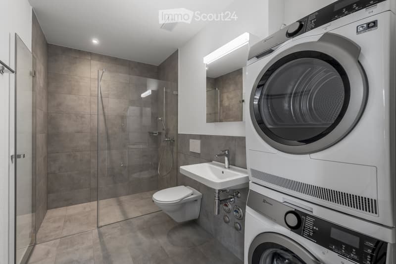 Modern bathroom with a toilet, sink, shower, and a stacked washer/dryer unit. Features include tiled walls, a glass shower enclosure, and built-in lighting.