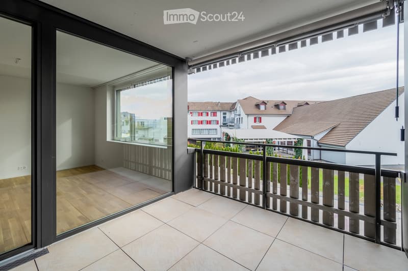 Balcony with tiled floor, wooden railing, large glass sliding doors leading to a room with wooden flooring, and a view of neighboring houses.