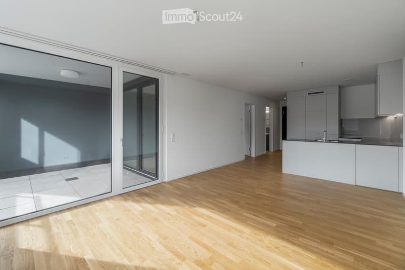Bright, empty room with wooden flooring, large glass sliding doors, and a modern kitchen area visible in the background.