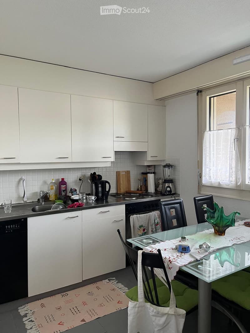 A modern kitchen with white cabinets, a sink, an oven, a dishwasher, and a dining area with a glass table and chairs.