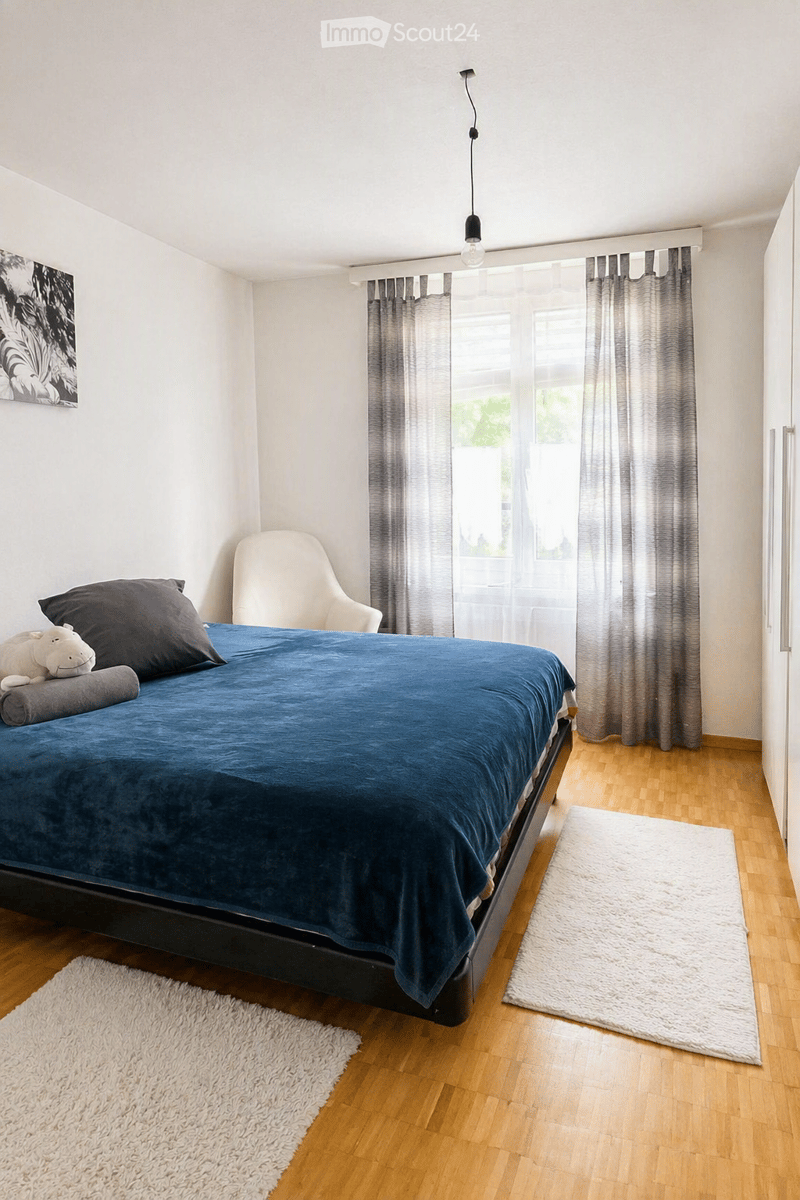 A modern bedroom with a large bed covered in a blue duvet, pillows, and a stuffed animal. A white armchair is positioned near the window, which has grey curtains. The room features light wooden flooring and two white rugs placed on either side of the bed.